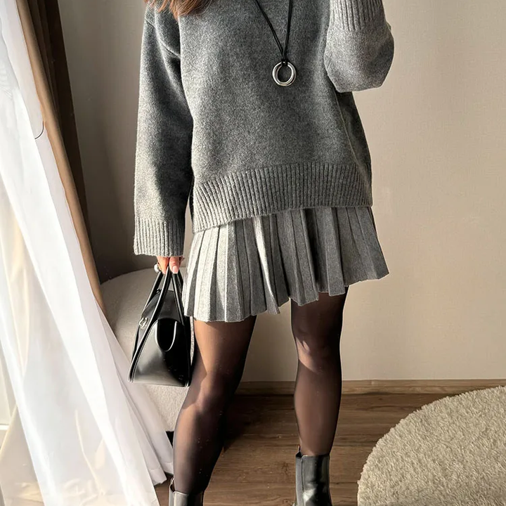 A person wearing a gray knit sweater and matching pleated mini skirt, styled with black tights, black ankle boots, a long black necklace with a silver ring pendant, and holding a black handbag in an indoor setting.