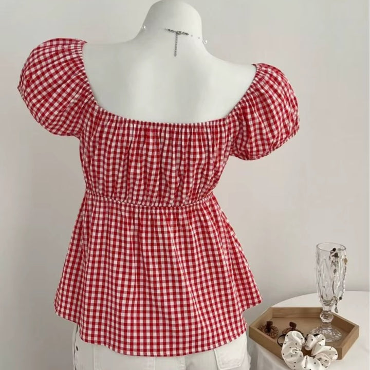 Red Gingham Babydoll Top – Cottagecore Aesthetic Blouse. Red and white gingham off-the-shoulder puff-sleeve blouse displayed on a white mannequin torso, with a small decorative tray and glass on a side table in the background.